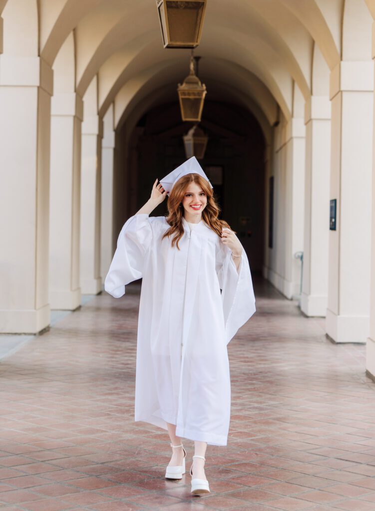 College graduate in an all white cap and gown walking the halls of UCLA for her graduation photos with Emily Winnie Photography.