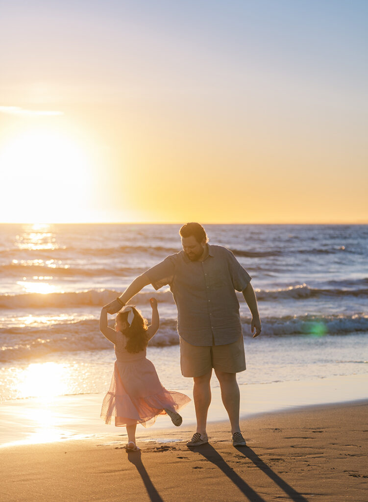 A dad dancing barefoot on the beach with his toddler daughter during a sunset session in Los Angeles.